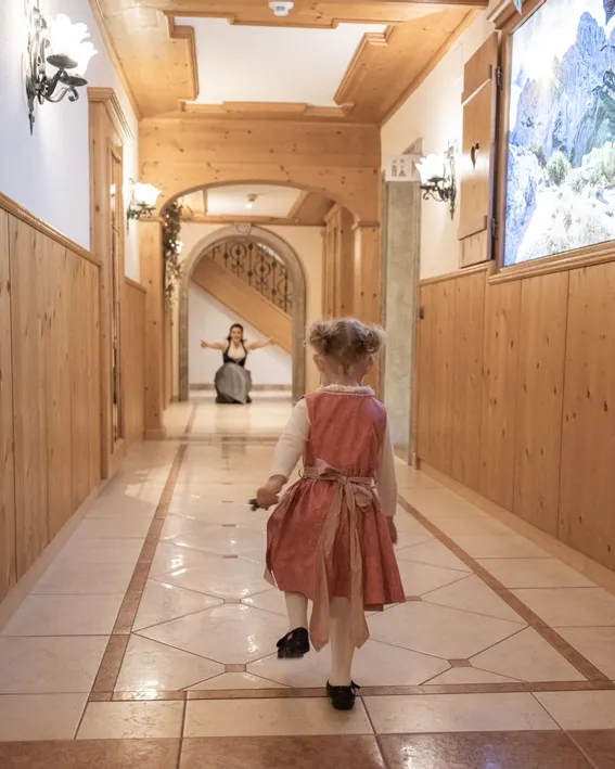 Kleines Mädchen im Dirndl geht den Flur eines Alpenhotels zur Frau hinunter